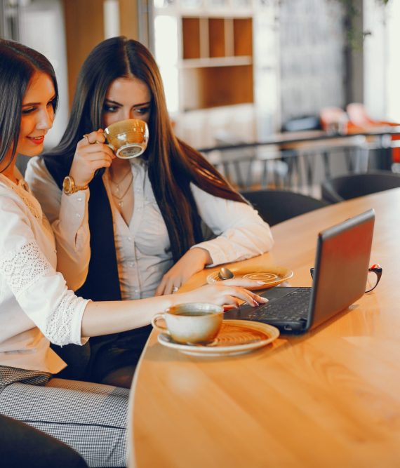 an elegant and stylish young lady with long dark hair dressed in a white shirt sitting in a restaurant with a beautiful interior with her young and beautiful girlfriend dressed in a white shirt and black jacket, they drink coffee together and using a laptop